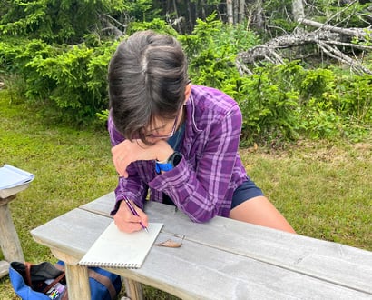 Person sitting outdoors at a picnic table writing in a notebook among trees.