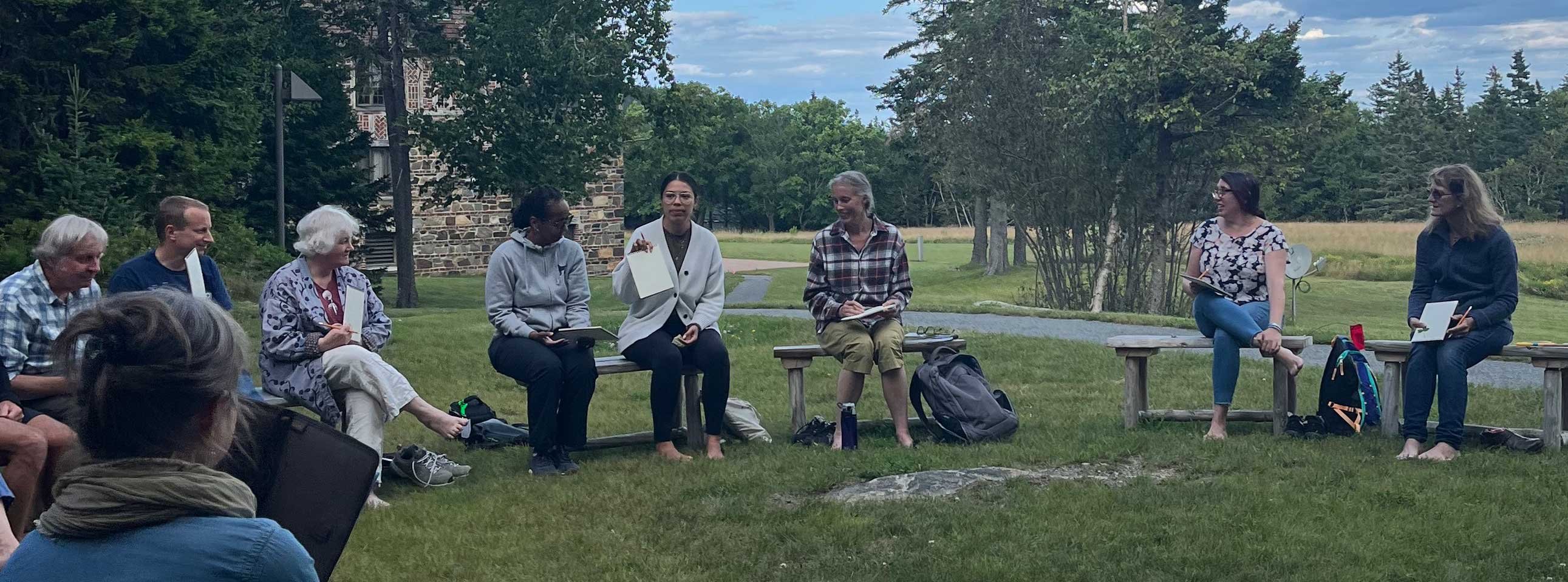 Group of people in a circle outside, discussing in a grassy area with trees.